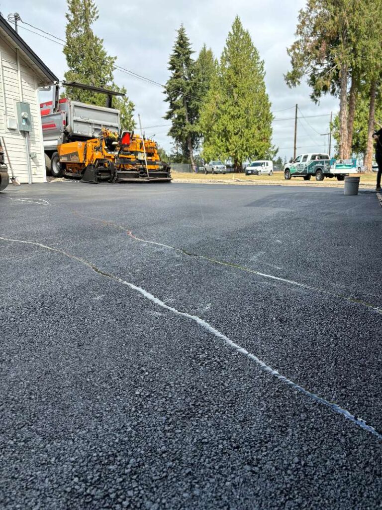 Asphalt surface with a sealed crack line in the foreground, with paving equipment and work trucks staged in the background.