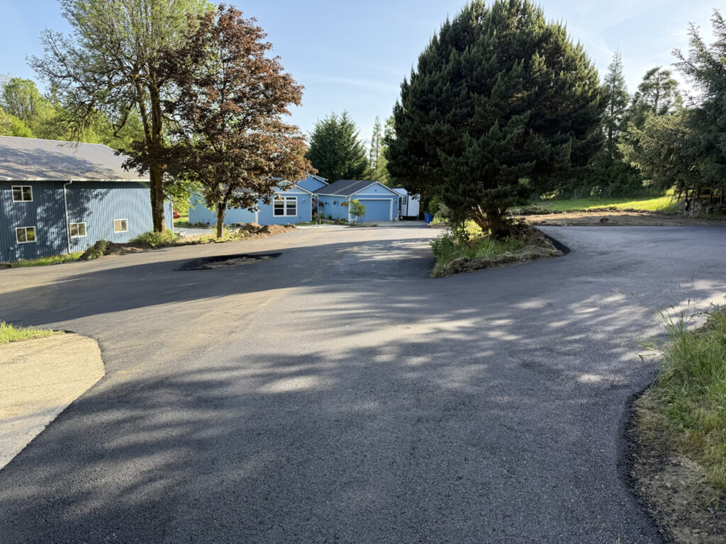 Newly paved asphalt driveway with a smooth, dark finish leading to blue buildings, surrounded by trees and landscaping.