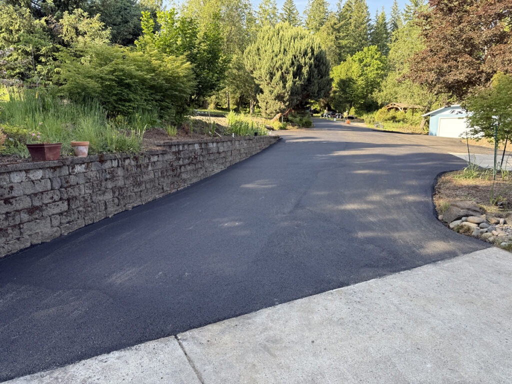 Newly paved asphalt driveway with a smooth black finish beside a stone retaining wall and landscaped trees.