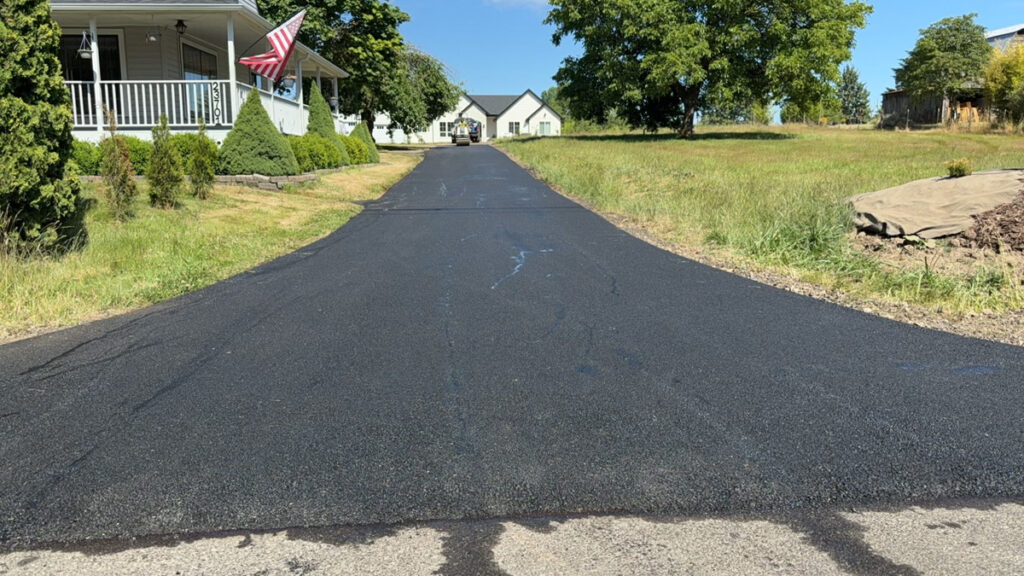 Long newly paved asphalt driveway with a smooth black surface leading to a home, bordered by grass and landscaping.