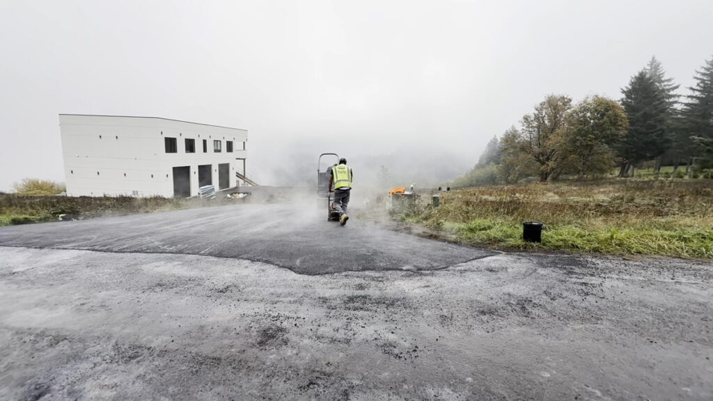 Worker applies asphalt during commercial paving services on a wide paved area near a commercial building in a misty outdoor setting.